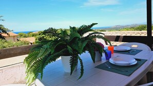 A table with a potted fern, plates, and cutlery on a terrace with a view of the sea and hills. - Case Vacanza Rena Majore (Aglientu)