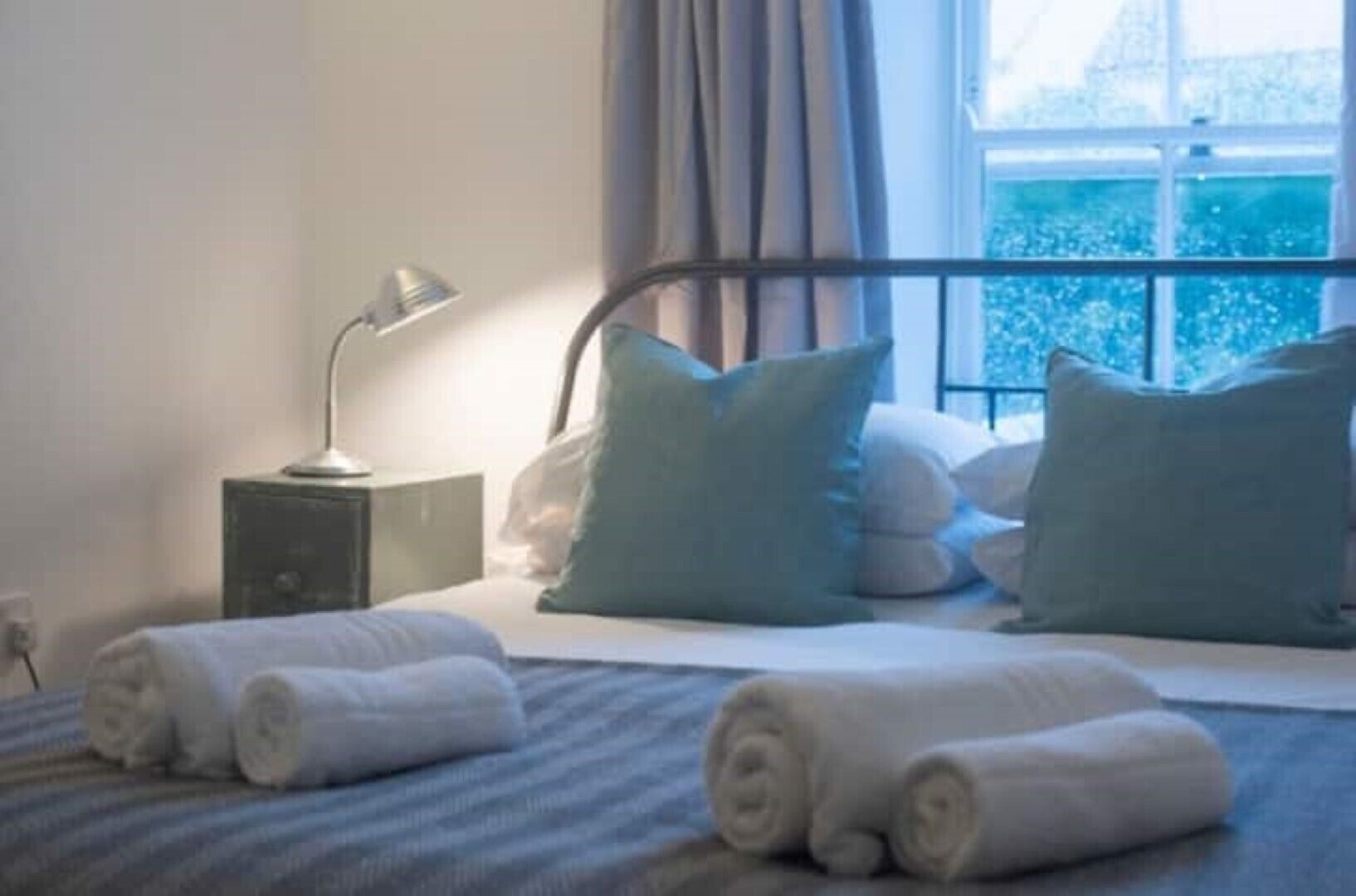 A neatly made bed with rolled towels, a bedside lamp, and pillows on a window seat with a view of a cityscape.