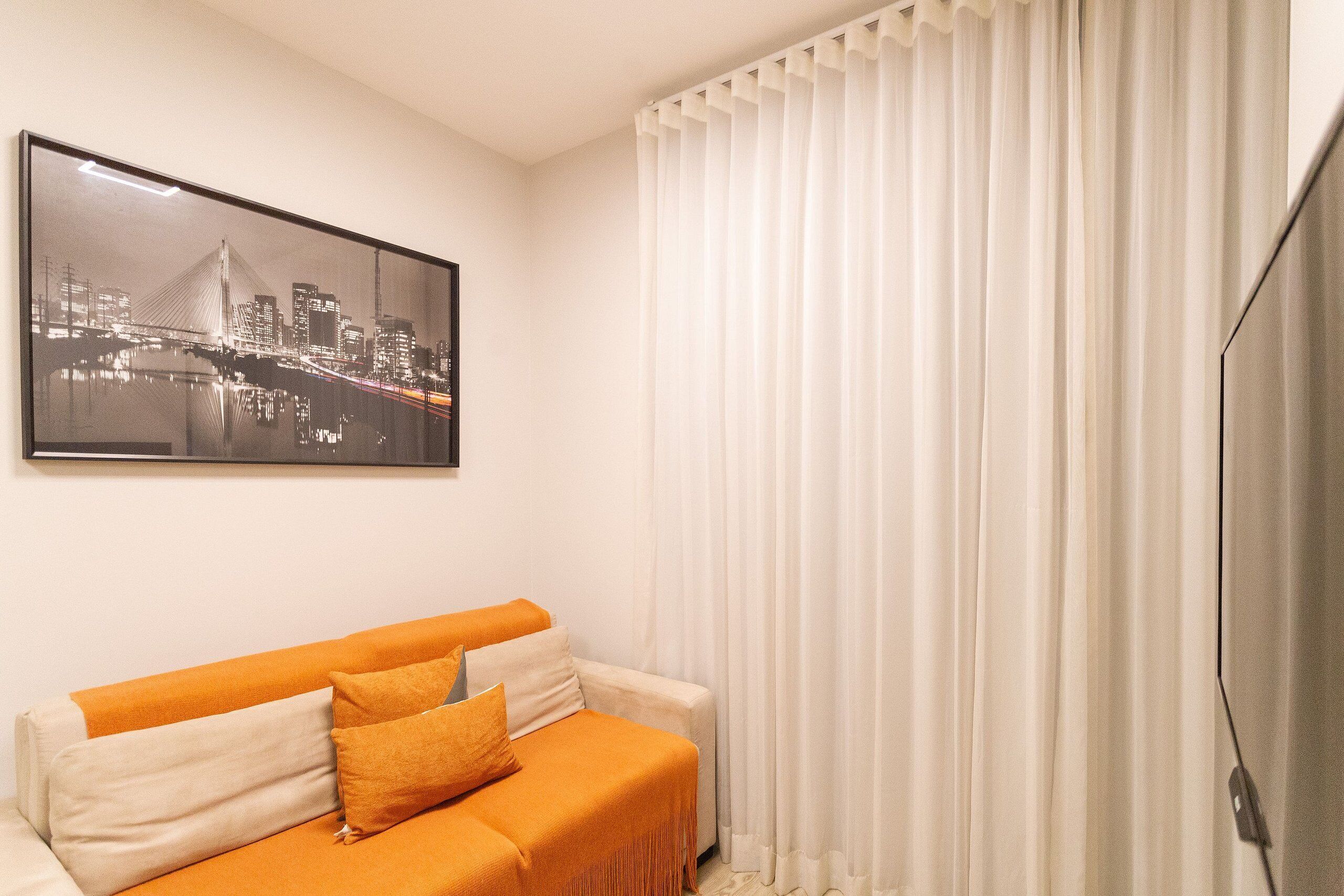 A living room with a sofa, a framed cityscape picture, and a large window with white curtains.