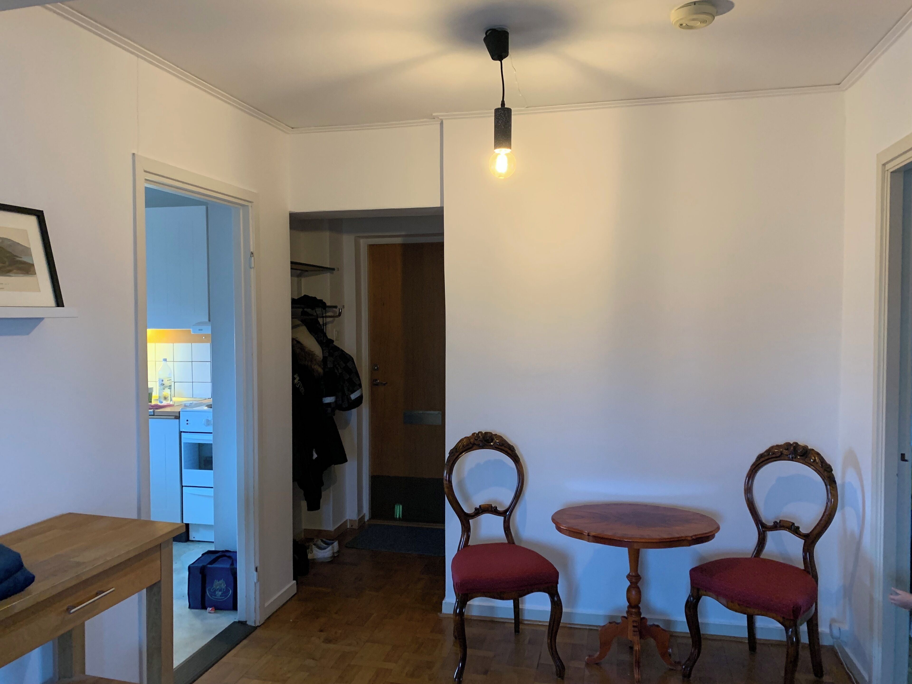A room with a wooden table and two chairs, a coat rack with a coat, and a door leading to another room.