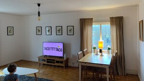 A living room with a TV displaying 'TACOT TACO', a dining table with chairs, a small white table, and framed pictures on the wall.