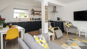 Una cucina moderna con mobili scuri, un piano di lavoro bianco e un forno integrato. C'è una zona pranzo con un tavolo bianco, sedie gialle e un divano grigio con cuscini decorati. Un televisore è montato a parete e una finestra offre vista su spazi verdi.