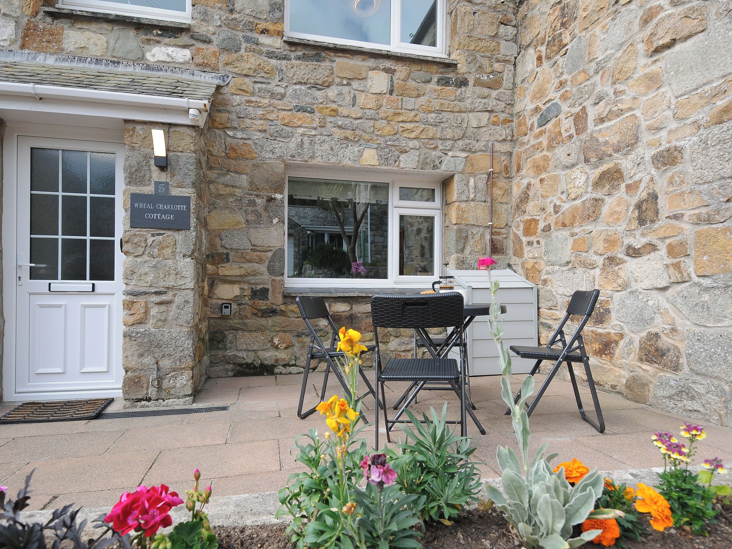 A stone cottage with a white door, a window, and a small outdoor seating area with two chairs and a table.