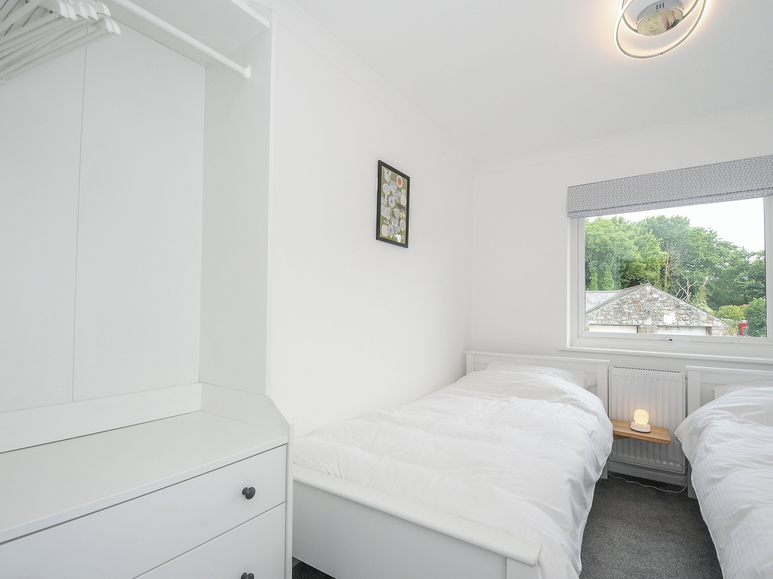 Ein Schlafzimmer mit zwei Betten, einer weißen Kommode, einem gerahmten Bild, einem Fenster mit Blick auf Grünflächen und einer wandmontierten Lampe.