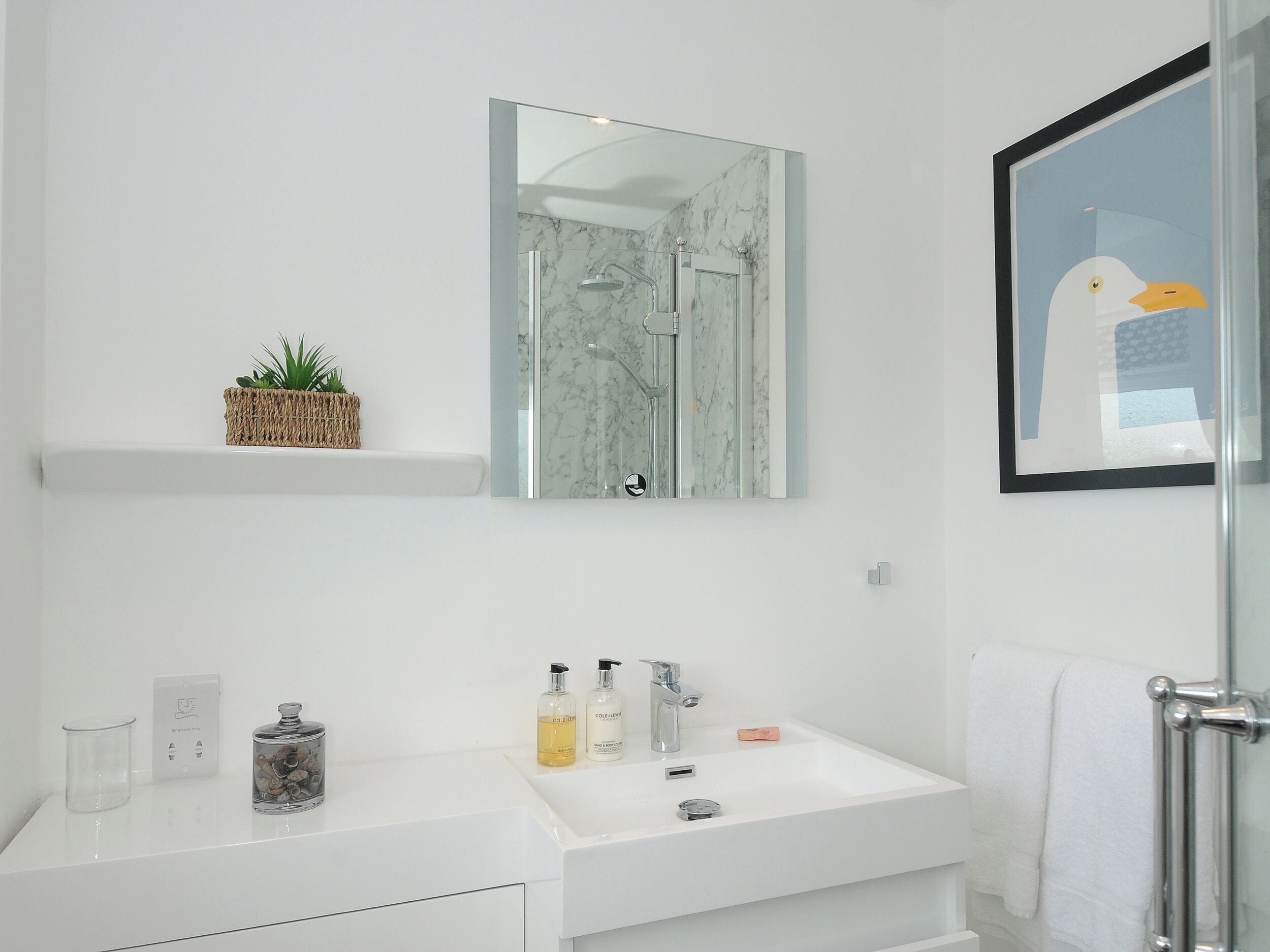 Un baño moderno con un tocador blanco, cabina de ducha con mampara de vidrio y una obra de arte con un dibujo de una gaviota enmarcada.