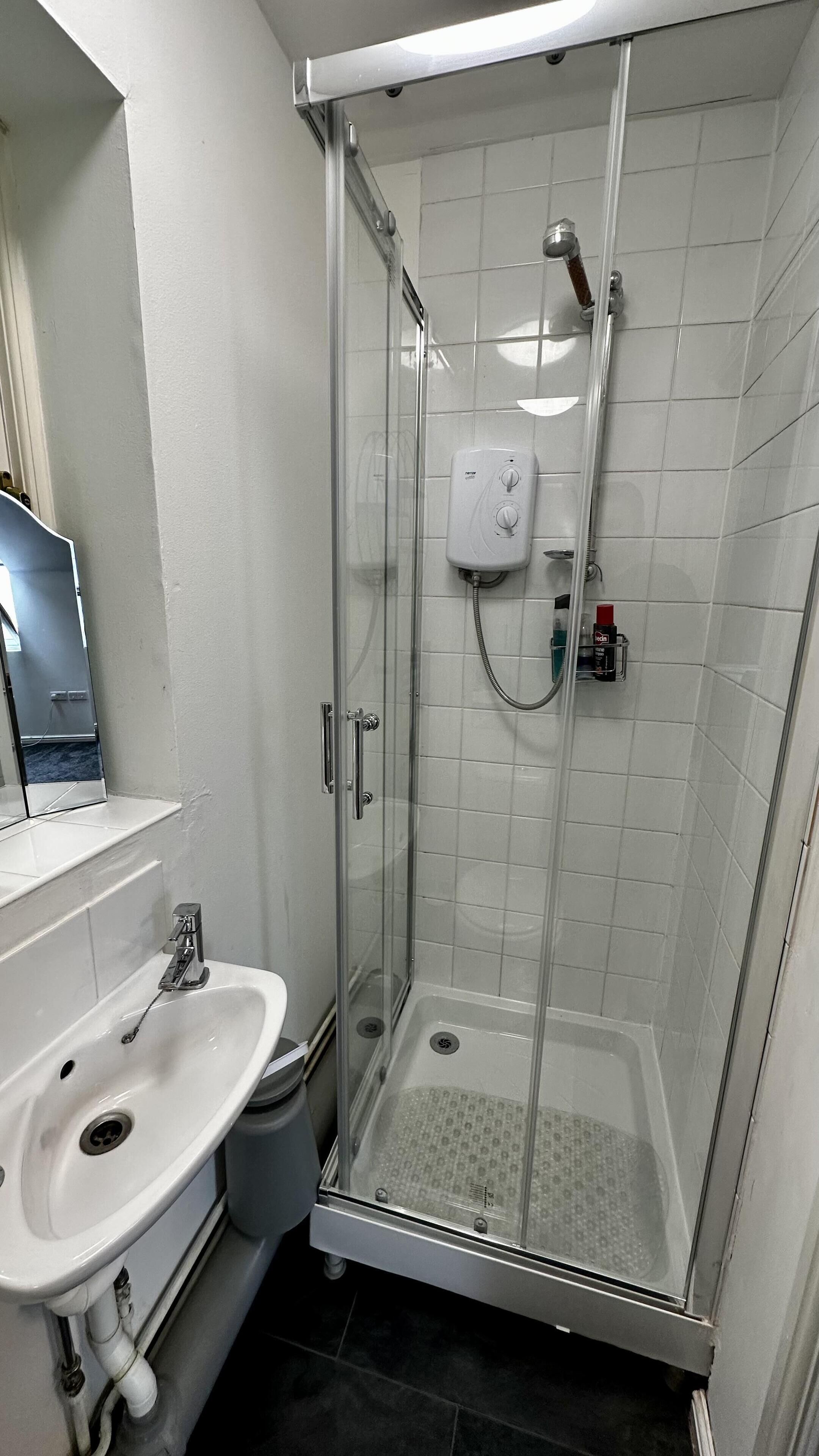 A glass-enclosed shower with a handheld showerhead, a handheld soap dispenser, and a small shelf with a bottle.
