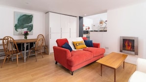 Una sala moderna con un sofá rojo, una mesa de comedor de madera y sillas. Se ve una chimenea y una cocina al fondo.