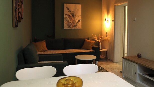 Een gezellige woonkamer met een donkergrijze bank, een kleine ronde tafel en een witte stoel. Op de tafel staat een decoratieve vaas met bloemen.
