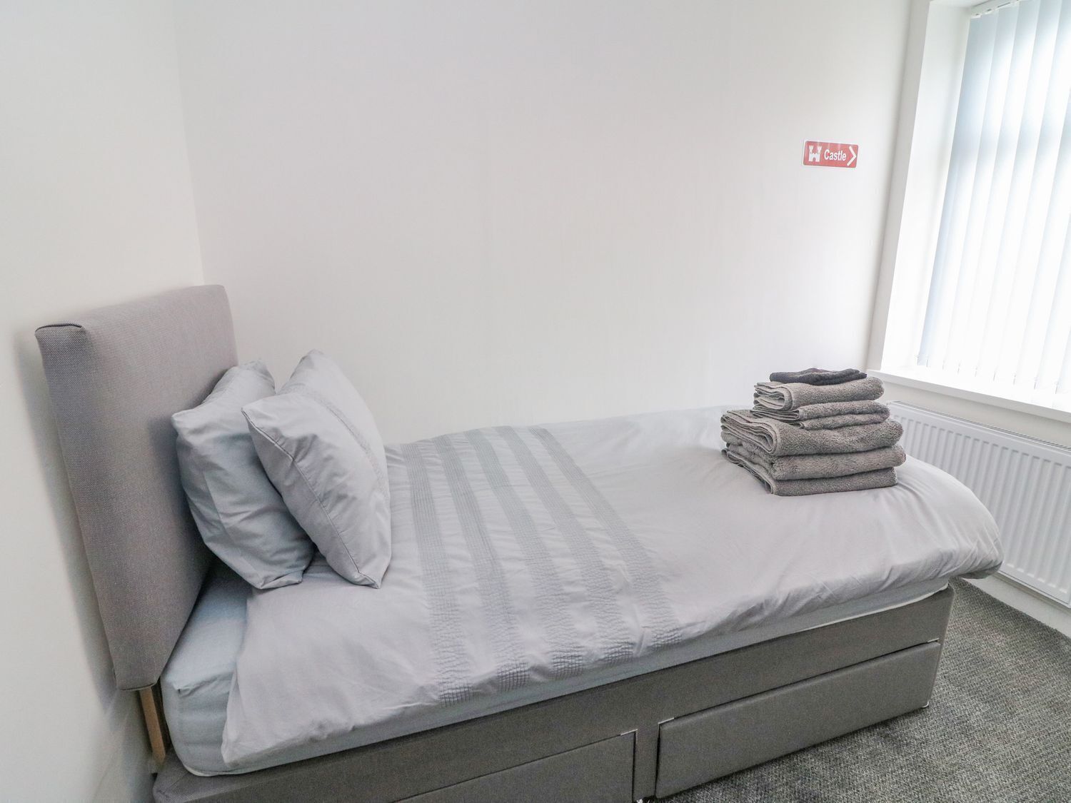 A single bed with a grey headboard and pillows, a folded grey blanket, and a radiator in the background.