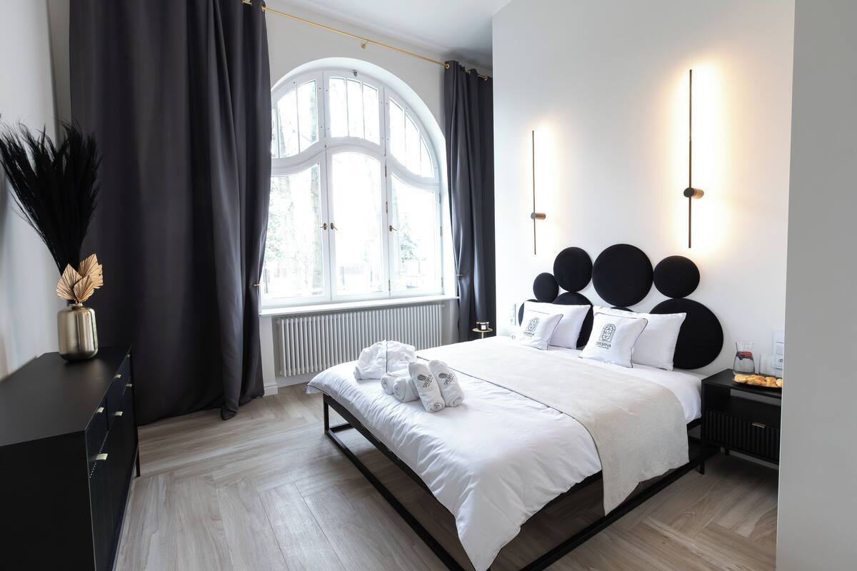 Une chambre d’hôtel moderne avec une grande fenêtre, un lit avec du linge de lit blanc, une table de chevet noire et un luminaire fixé au mur.