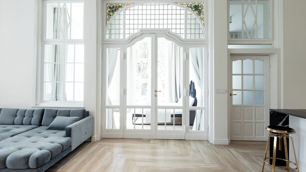 A spacious living room with a large grey sectional sofa, a wooden floor, and a glass door leading to a balcony. - Srebrna Luxury Apartments (Lodz)