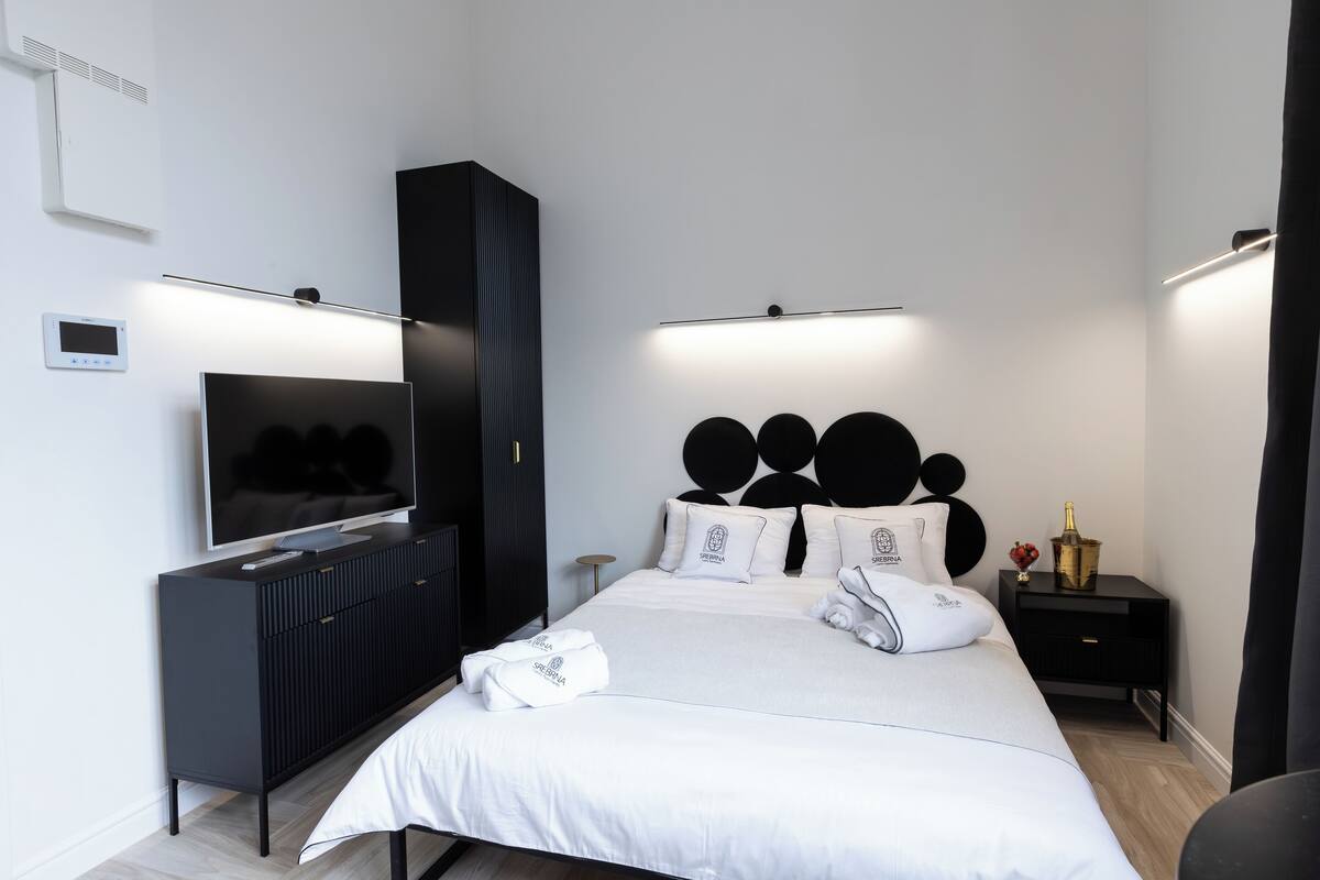 Une chambre d’hôtel moderne avec un grand lit, une télévision, une armoire noire et une table de chevet sur laquelle se trouvent une bouteille et des fleurs.