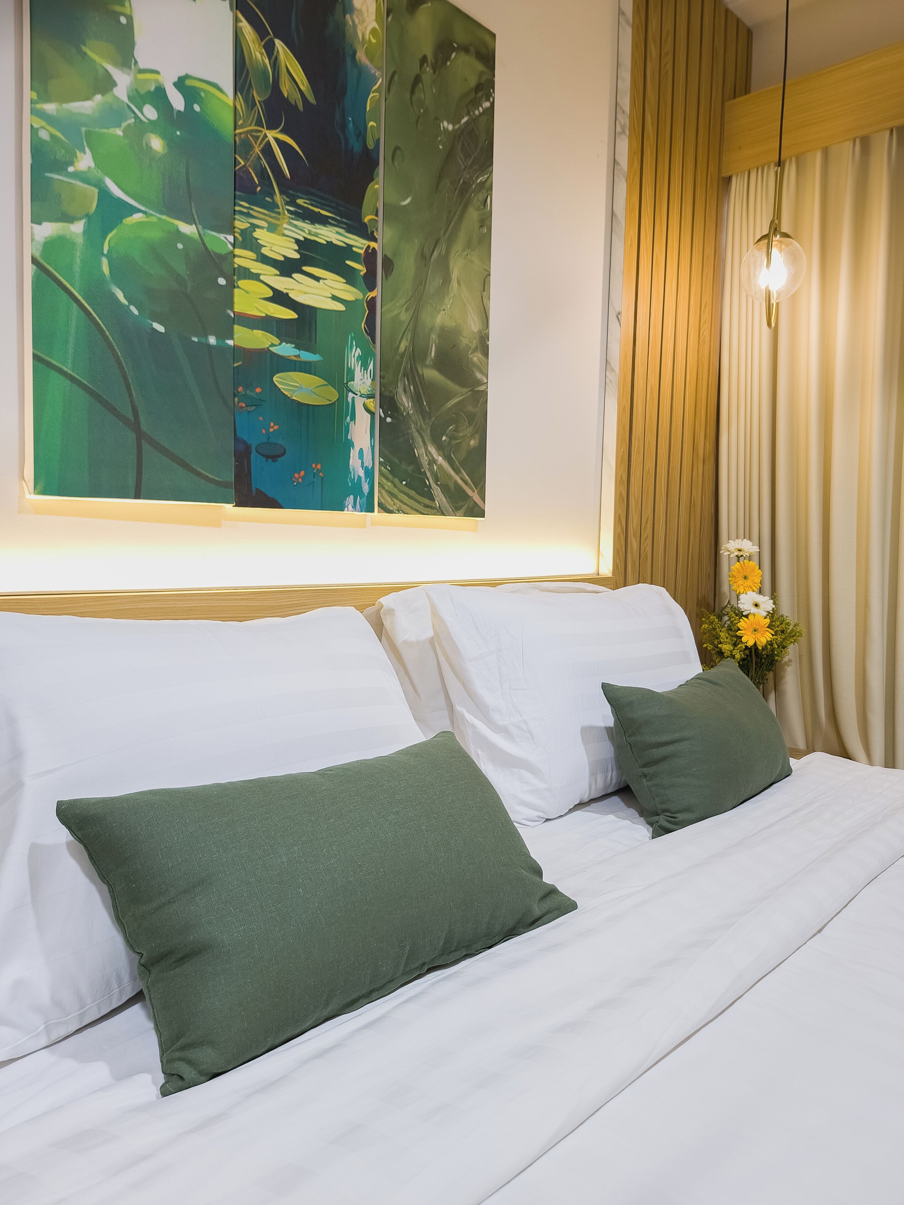 Una cama bien hecha con sábanas blancas y almohadas verdes, un cuadro abstracto grande en la pared y un jarrón con flores en la mesita de noche.