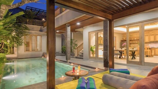Een zwembadterras met een houten pergola, een tafel met handdoeken en uitzicht op het zwembad en het aangrenzende gebouw.