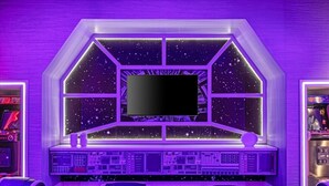 Habitació amb un gran pantalla de televisió, consoles de joc i diverses butaques gamer.