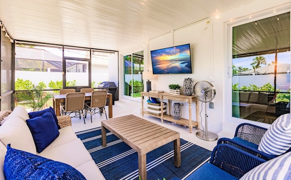 A spacious patio with a wooden table, wicker furniture, and a large TV screen displaying a sunset.