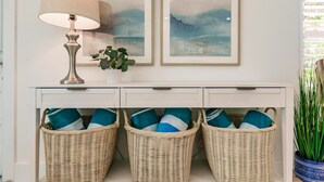 A white console table with three wicker baskets containing blue and white pillows, a lamp, and two framed abstract paintings. - Chic Bungalow with Fire Pit, Lanai & Games! (Naples)