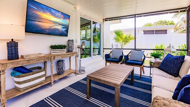 A sunlit room with a wooden table, two blue chairs, a sofa, and a large TV on the wall. - Chic Bungalow with Fire Pit, Lanai & Games! (Naples)