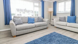 A modern living room with two grey sofas, a blue rug, and large windows with blue curtains. - Cynefin (Llangefni)