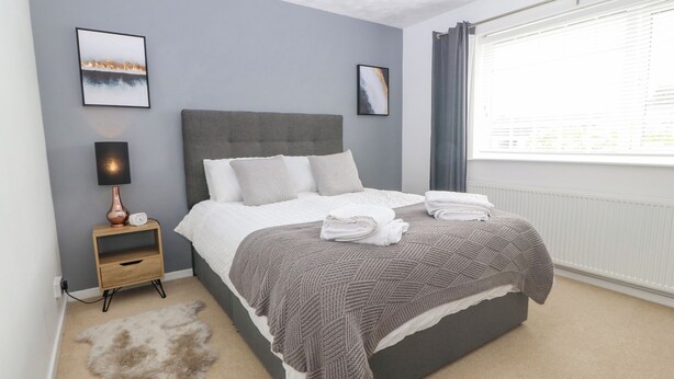 A modern bedroom with a grey headboard, a bedside table with a lamp, a fluffy rug, and a window with curtains. - Cynefin (Llangefni)