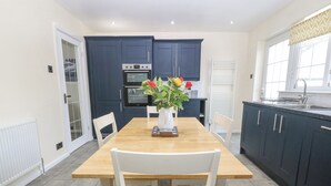 A modern kitchen with dark blue cabinets, a wooden dining table, and white chairs. - Cynefin (Llangefni)