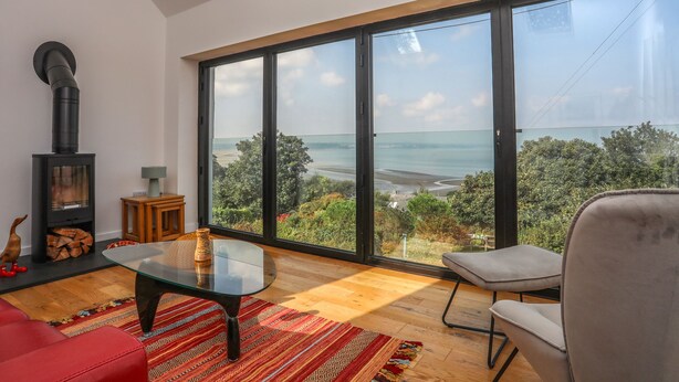 A modern living room with a large glass window, a fireplace, and a view of a beach. - Gorphwysfa (Beaumaris)