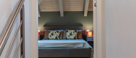 A bedroom with a bed, wooden flooring, a sloped ceiling, and a nightstand with a lamp.