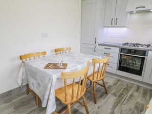A small, clean kitchen with white cabinets, a gas stove, and a dining table with chairs. - Maghera Caves Cottage (Ardara)