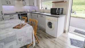 A compact kitchen with white cabinets, a washing machine, a microwave, and a dining area with a table and chairs. - Maghera Caves Cottage (Ardara)