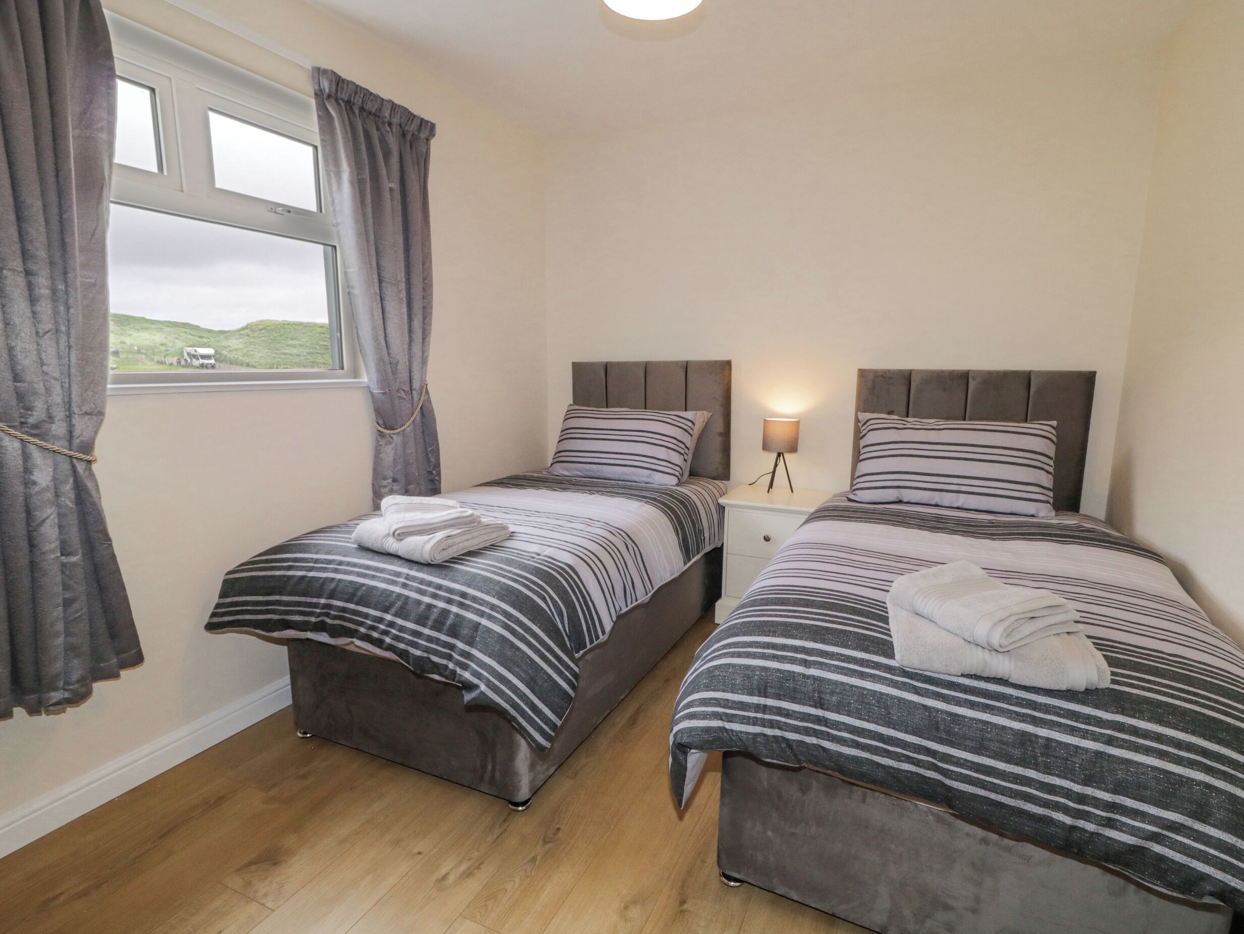 A bedroom with two beds, each with striped bedding and pillows, a bedside table with a lamp, and a window with curtains.