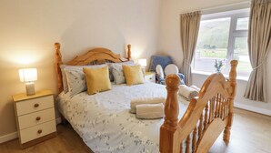 A bedroom with a wooden bed frame, a nightstand with a lamp, a chair, and a window with curtains. - Maghera Caves Cottage (Ardara)