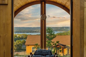 A view through an arched window with a clock, overlooking a lake, trees, and buildings.