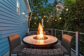 Una chimenea redonda al aire libre con fuego, rodeada de sillones y un sofá de mimbre en una terraza con barandilla.
