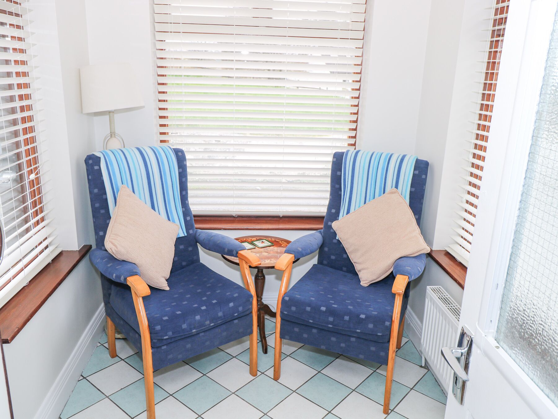 A corner room with two blue armchairs, a small wooden table, and a window with blinds.