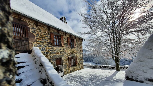 Un bâtiment en pierre, avec un toit et des fenêtres couverts de neige, entouré d’arbres et d’un sol également recouverts de neige.