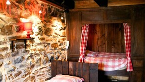 Une chambre rustique avec un mur en pierre, un lit en bois surmonté d’un baldaquin à carreaux et une petite table avec une lampe rouge.