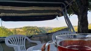 Une table avec une chaise en plastique blanche et un verre de th é, placée sous un auvent rayé, avec en arrière-plan une vue sur des collines verdoyantes.