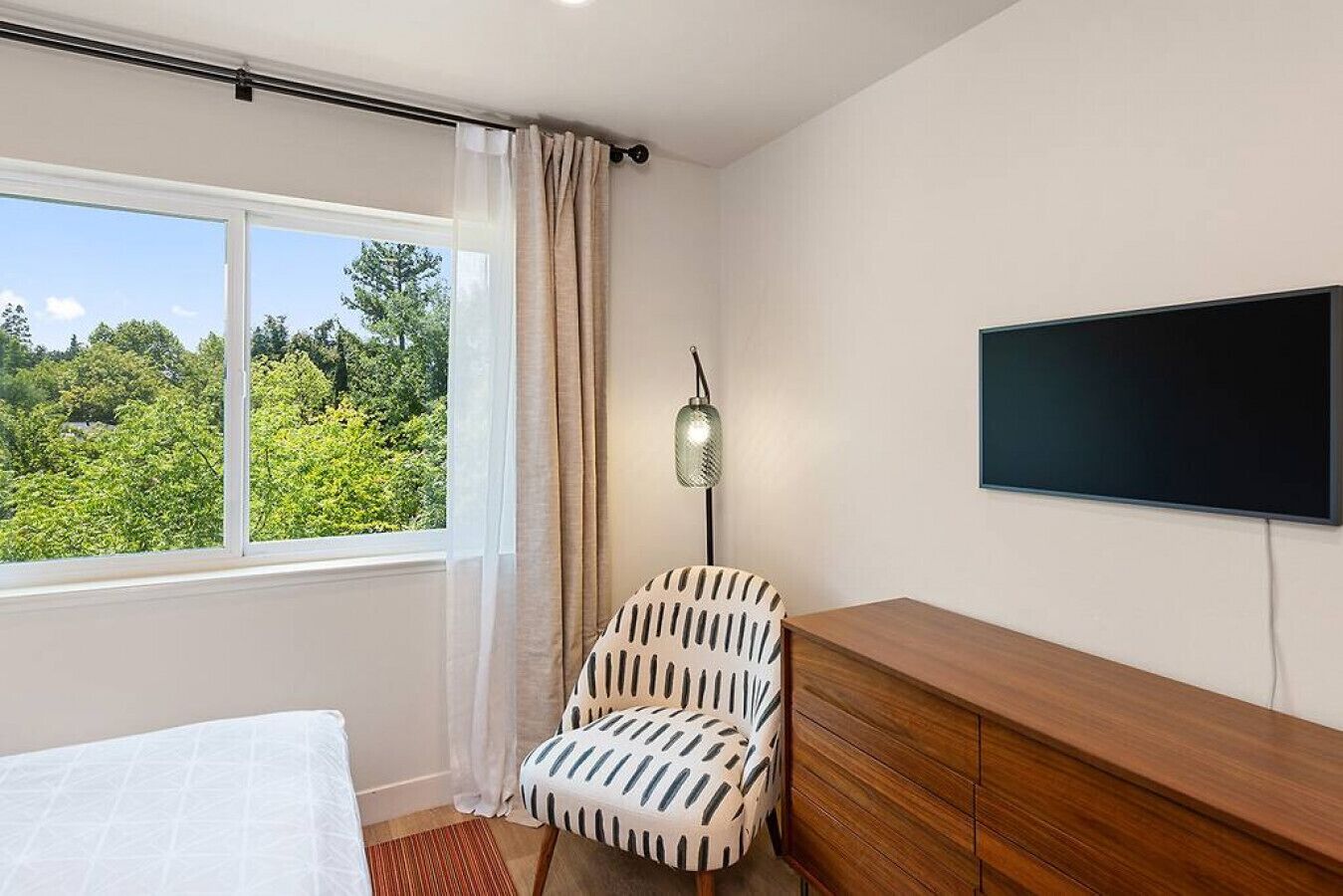 A bedroom with a bed, a chair, a TV, and a window with a view of greenery.