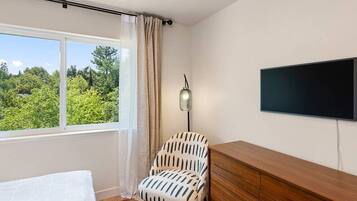 A bedroom with a bed, a chair, a TV, and a window with a view of greenery.