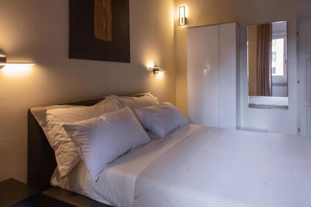 A neatly made bed with white linens and pillows, a dark headboard, and two wall-mounted light fixtures.