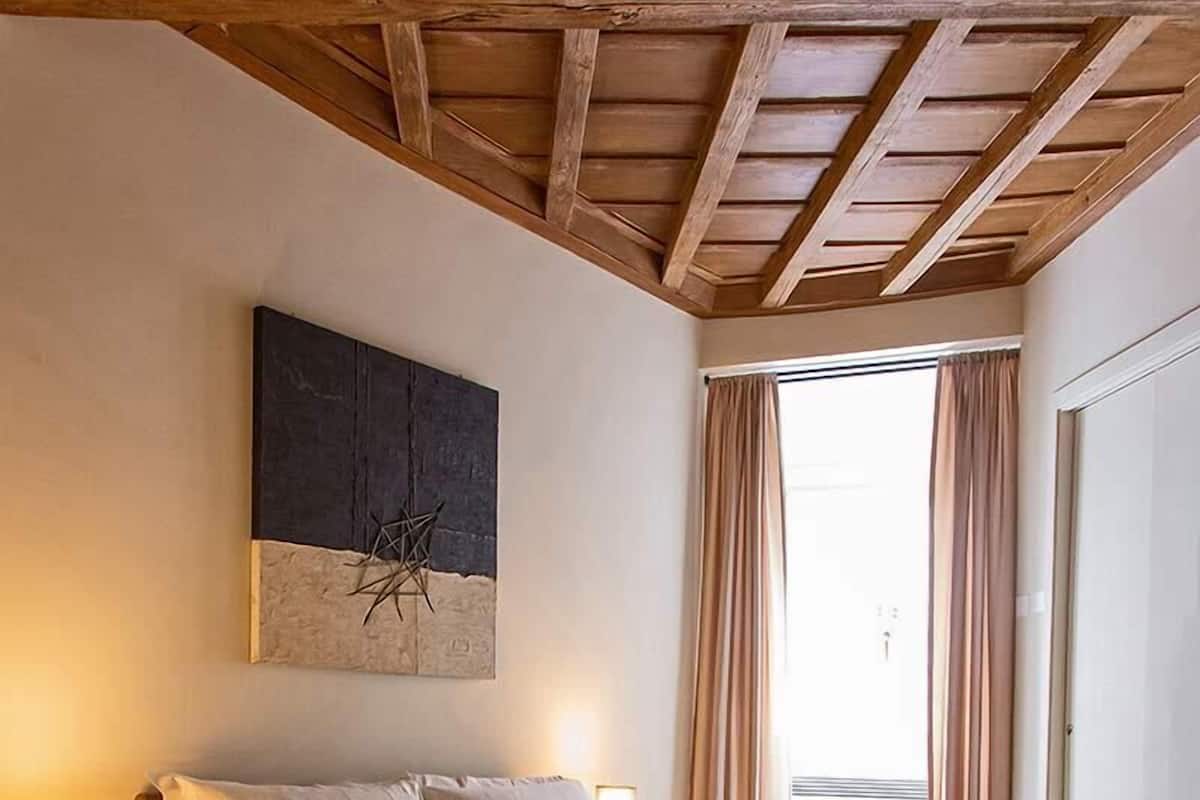 A bedroom with a wooden ceiling, a bed with white linens, a nightstand with a lamp, and a large window with curtains.