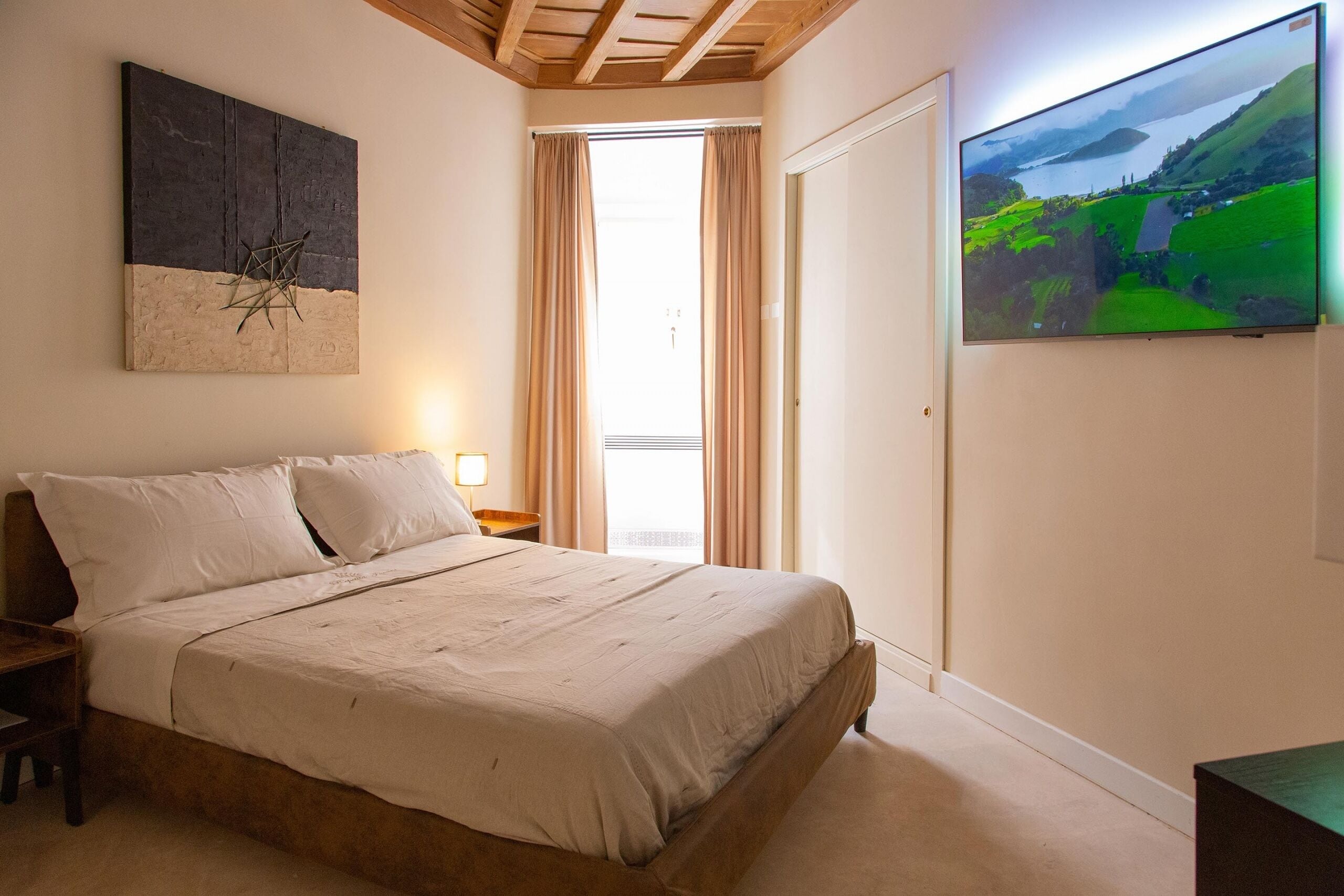 A bedroom with a bed, a painting, a TV, and a window with curtains.