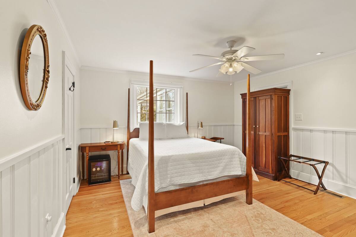 A bedroom with a four-poster bed, a ceiling fan, a wooden wardrobe, and a fireplace.
