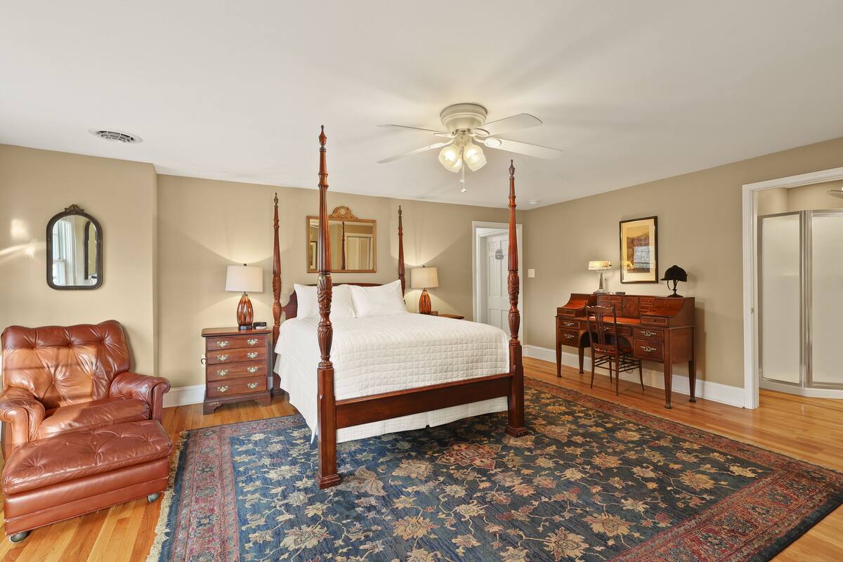 A bedroom with a four-poster bed, a desk with a chair, a leather armchair, and a large rug.