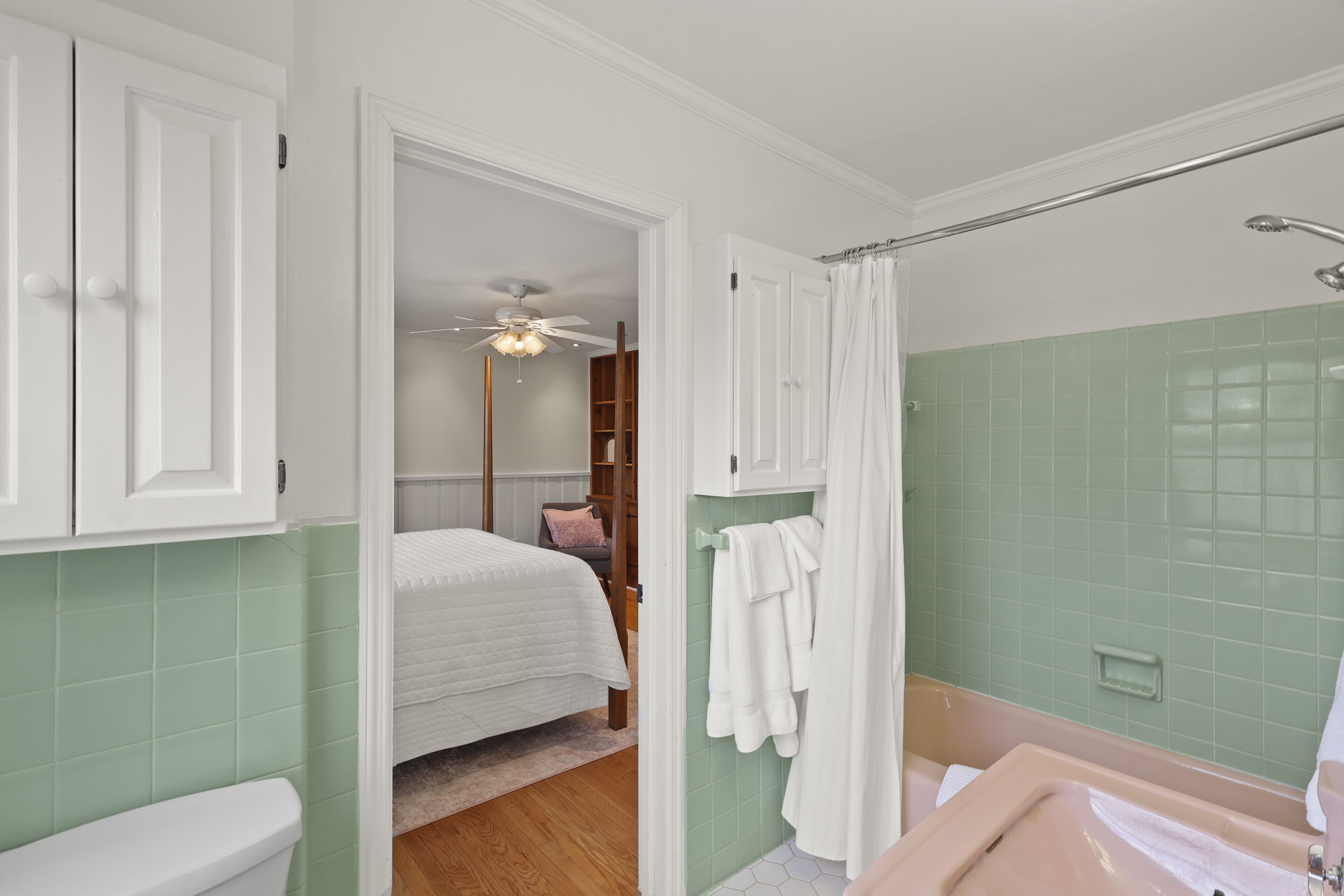 A bathroom with a toilet, sink, and shower. There is a bed in the adjacent room.