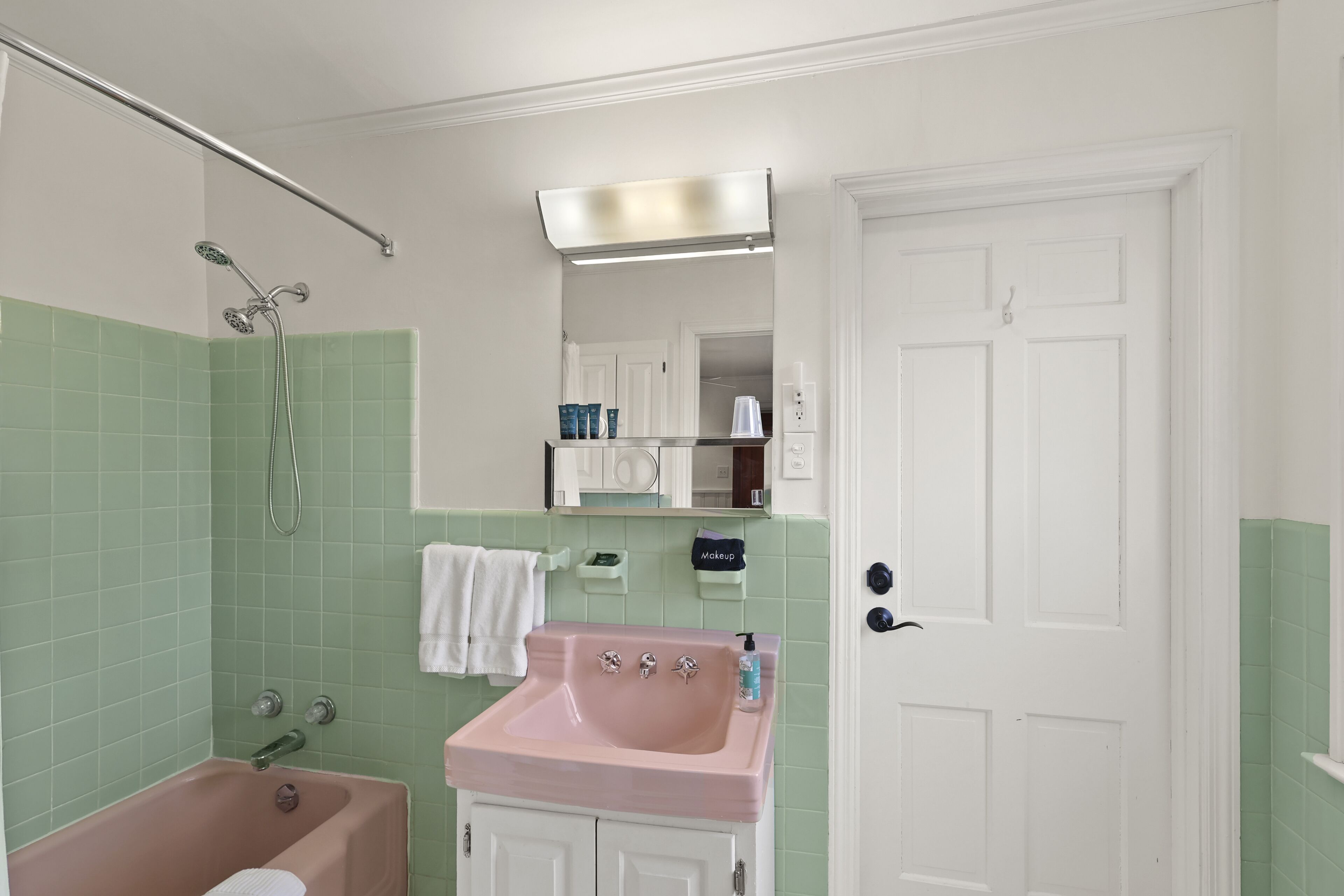 Un baño con un lavabo rosa, azulejos verdes, una puerta blanca y un espejo sobre el lavabo.