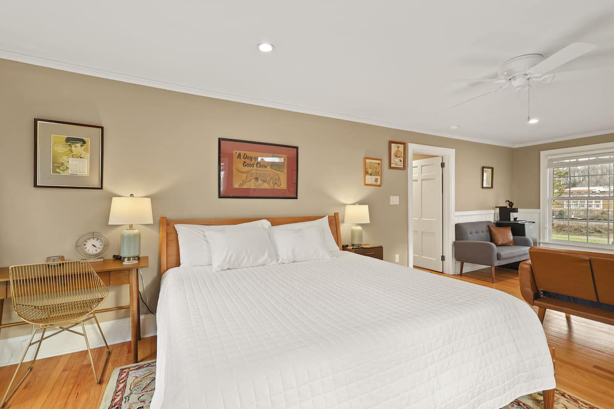 A bedroom with a large bed, wooden headboard, two bedside tables with lamps, a chair, a sofa, and a window with a view.