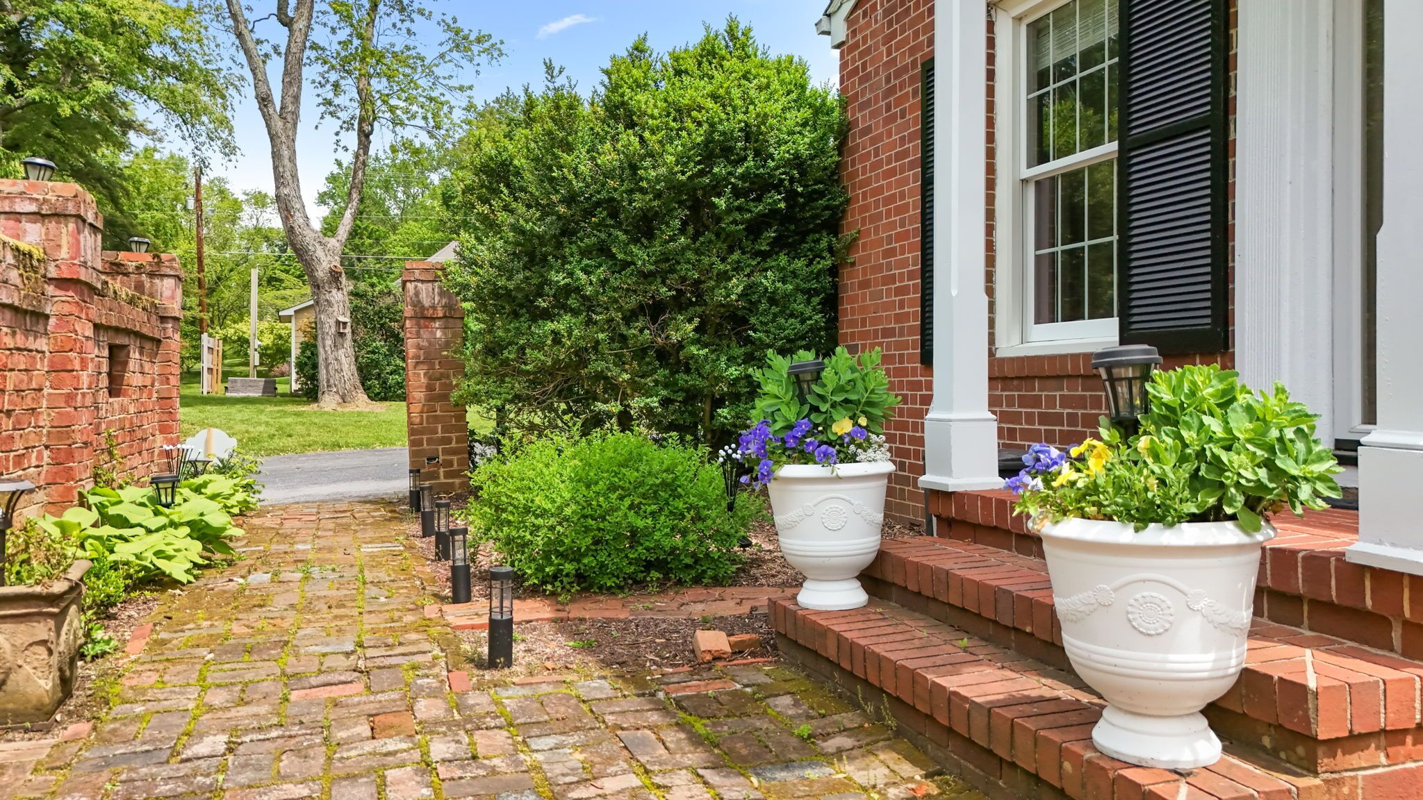 A brick house with a paved walkway, large potted plants, and a well-maintained garden.