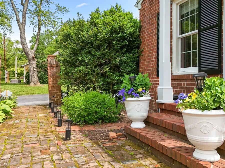 A brick house with a paved walkway, large potted plants, and a well-maintained garden.