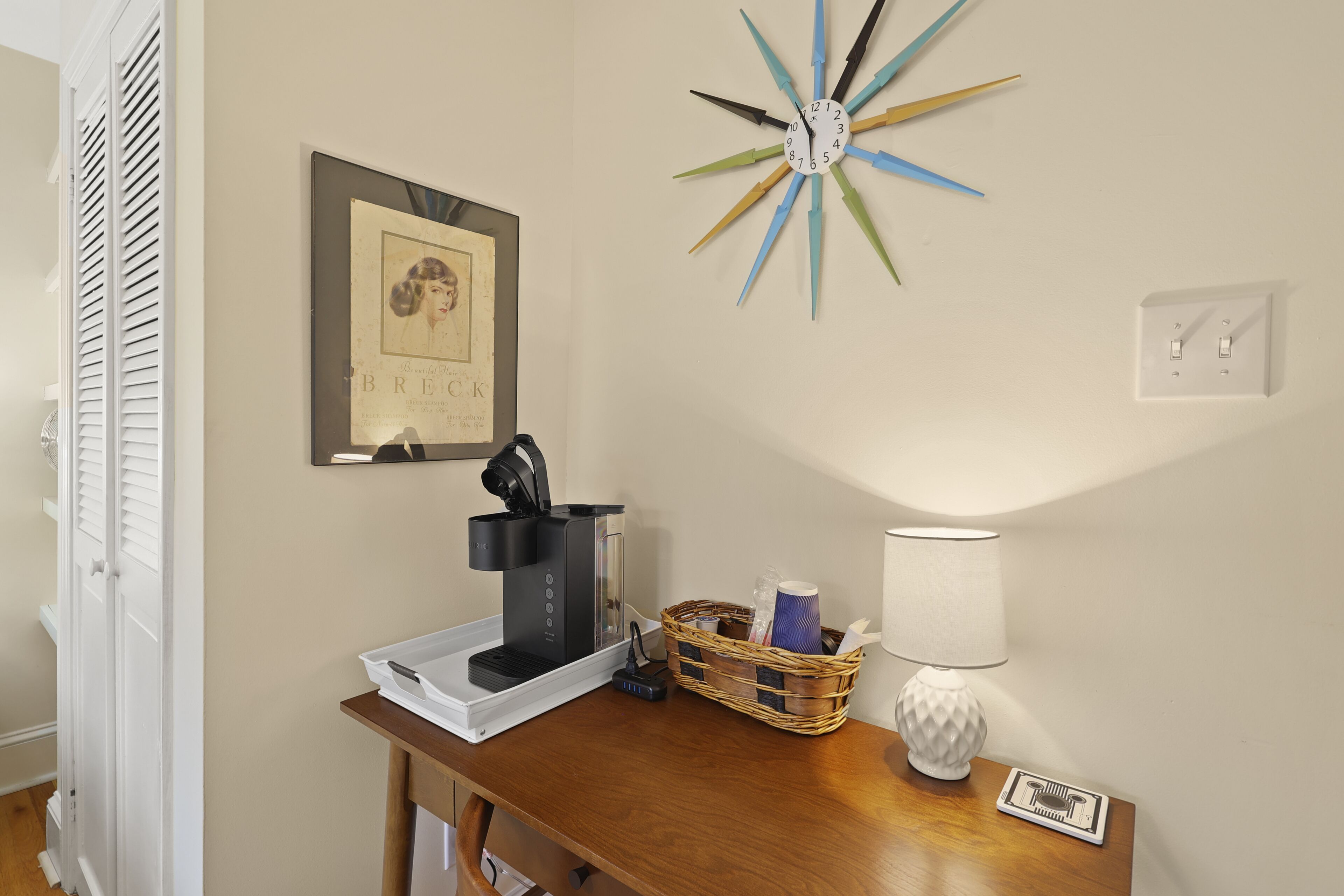 A room with a coffee maker on a wooden table, a framed picture on the wall, a star-shaped clock, and a lamp.